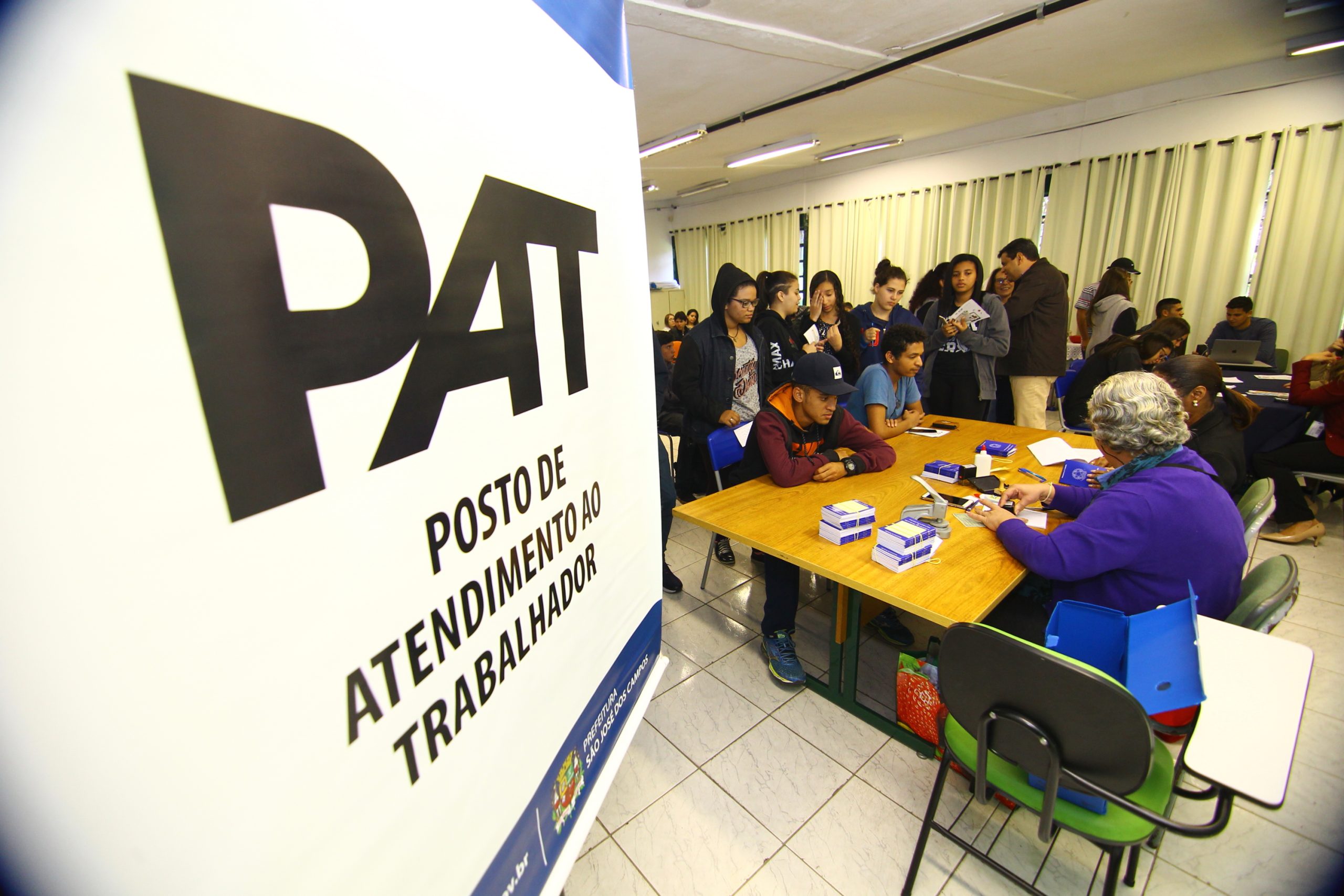 PAT de SJC, cidade que está em quinto lugar no ranking das que mais gerou emprego no estado de SP; na imagem jovens estão na fila com carteira de trabalho em mãos, aguardando para atendimento em frente à mesa com duas atendentes