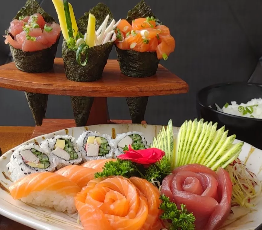 Prato com sashimis de salmão e atum, niguiri e sushis. Atrás um suporte de madeira com três tipos de temakis