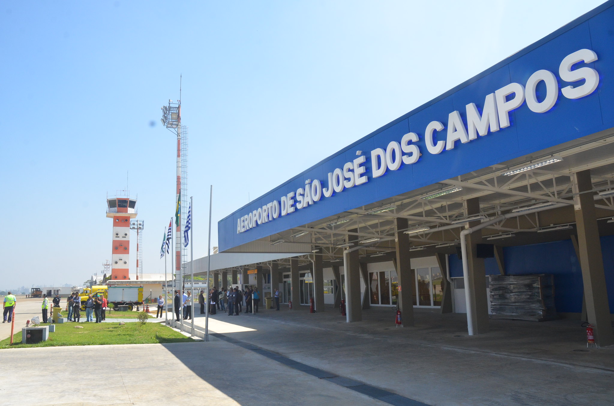 Fachada do Aeroporto de SJC, com céu azul, placa, área verde e hall na frente.