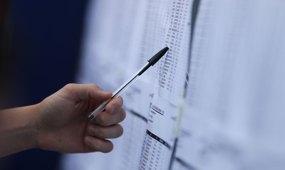 uma mão com uma caneta apontando para listas com nomes em papel sulfite colado na parede; alunos que fizeram Enem podem se inscrever para o Sisu a partir desta segunda-feira (19 de junho)
