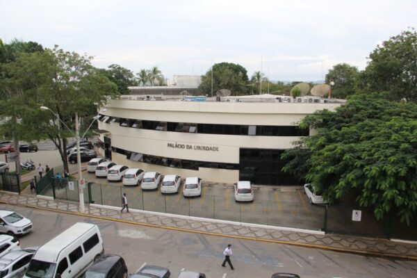 Vista aérea da Câmara de Jacareí, em meio às árvores, com céu branco, carros estacionados na frente e pessoas andando; foto ilustra matéria sobre reajuste salarial dos servidores municipais.