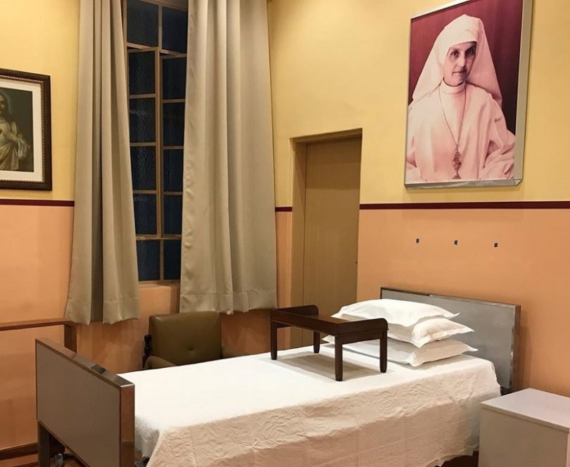 Quarto em que Madre Maria Teresa viveu, com uma cama, travesseiros, um quadro da madre na parede, uma janela e uma porta.