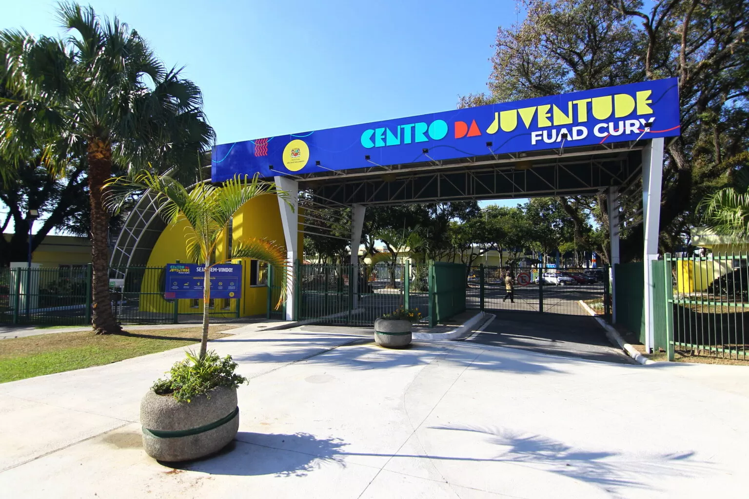 Fachada do Centro da Juventude, em São José dos Campos, que receberá feira noturna; foto mostra céu azul, placas em azul paredes amarelas e árvores.