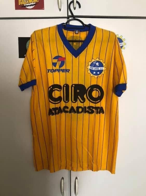 camisa amarela com listras, gola e manga nas cores azuis. Há também o escudo do São José e os patrocínios da Topper e do Ciro Atacadista