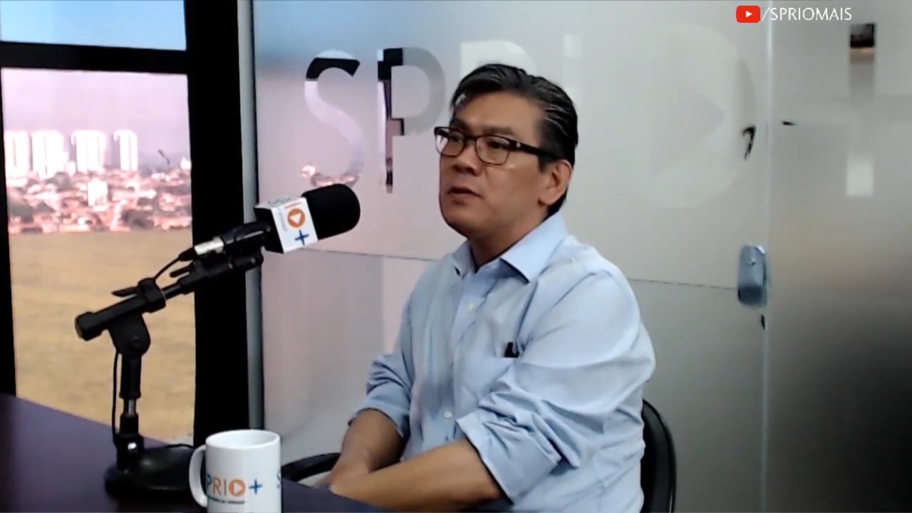 Dr. Ricardo Nakagawa em entrevista à SP RIO+ em que avaliou SUS e criticou mudança na taxação de planos de saúde. Ele está sentado, com a redação do portal desfocada ao fundo. Ele veste uma camis social azul, usa óculos preto e parece sério.