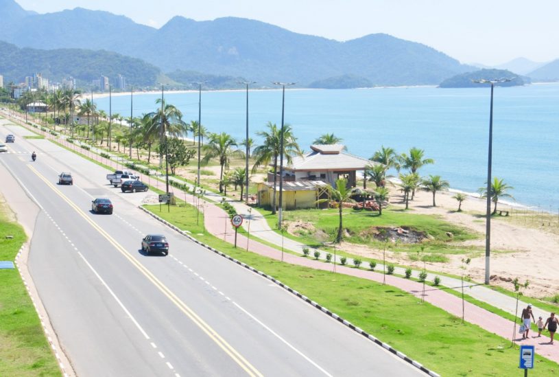 Foto aérea de rua da orla da praia, com um quiosque à esquerda e carros passando na rua.
