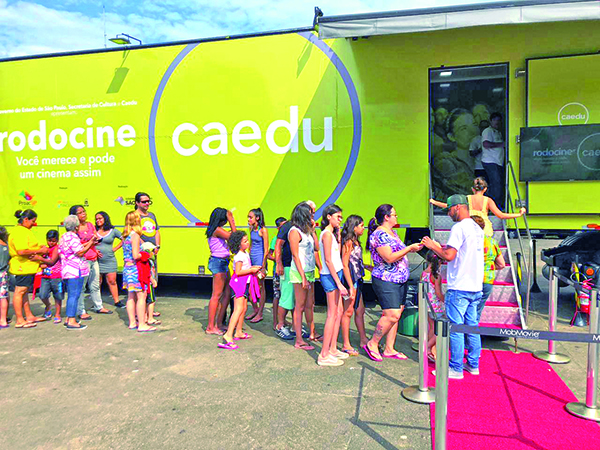 Na foto é possível ver a estrutura amarela do cinema móvel em um dia de sol e com o público em fila aguardando para entrar no local. Há um tapete vermelho esticado ao lado direito da foto. Ele leva à uma escada, onde se passa pela porta de acesso ao cinema.