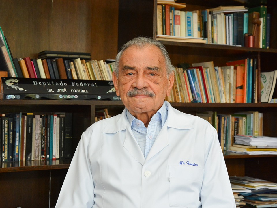 Dr. Coimbra, defensor da ozonioterapia, de jaleco médico e sentado em escritório com grande estante de livros atrás.