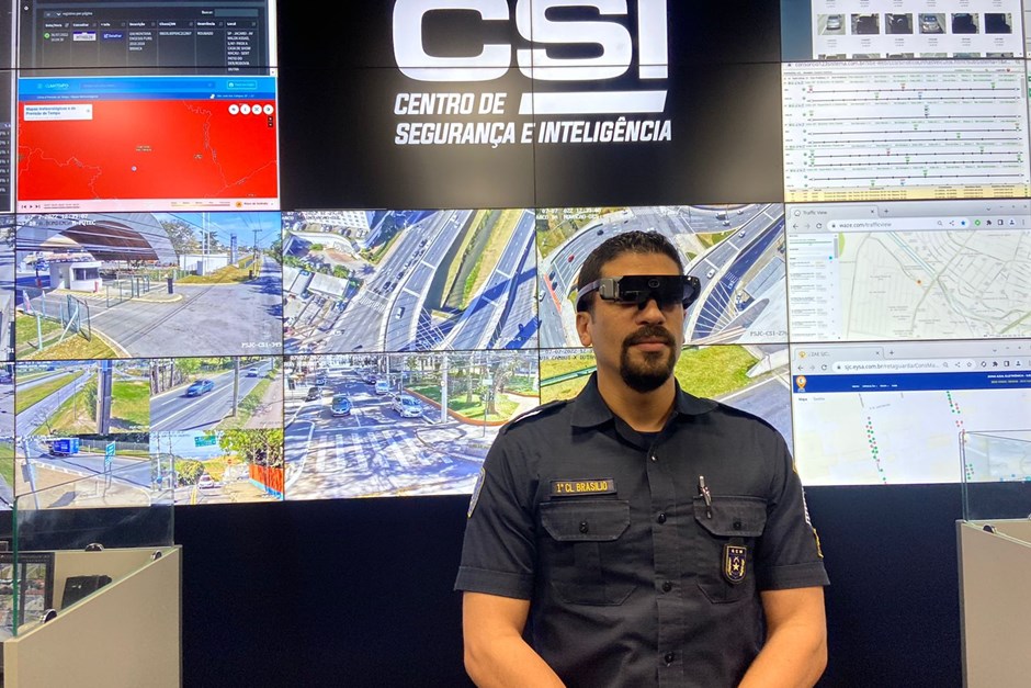 A foto mostra um agente de segurança fardado utilizando os óculos de alta tecnologia em frente ao painel do Centro de Segurança e Inteligência de São José dos Campos.
