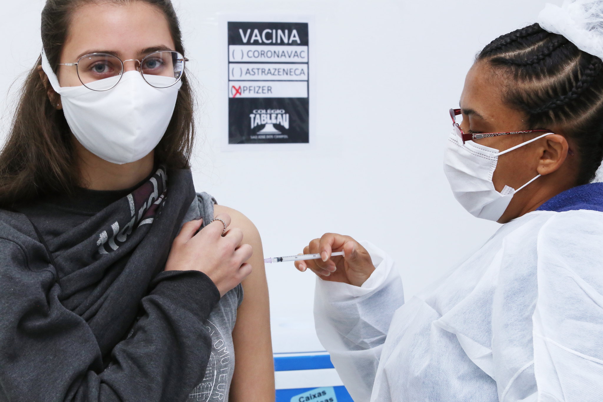 Jovem branca de olhos verdes e cabelo castanho recebendo vacina contra covid-19 de profissional da saúde negra com cabelo trançado e preso. A jovem usa óculos e máscara branca assim como a profissional, que ainda também está vestida com um jaleco branco. Ao fundo, se vê uma placa na parede com os nomes das vacinas Coronavac, Astrazeneca e Pfizer, esta última com um "x" vermelho marcado ao lado esquerdo.
