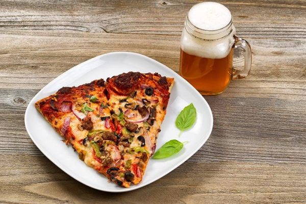 Um pedaço grande de pizza sob um prato branco ao lado de uma caneca com cerveja.