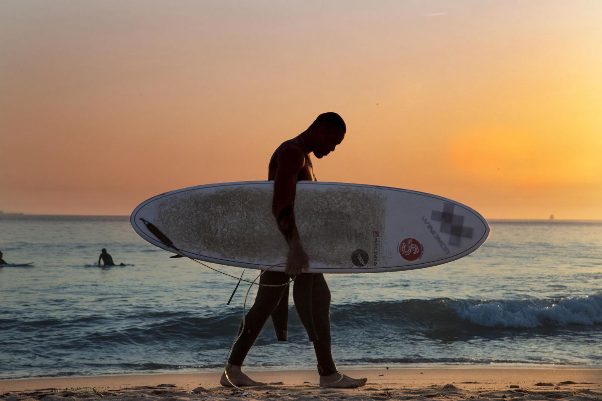 Homem com uma prancha nos braços, caminhando sobre a praia, simulando o Cine Outdoor Experience 2022.