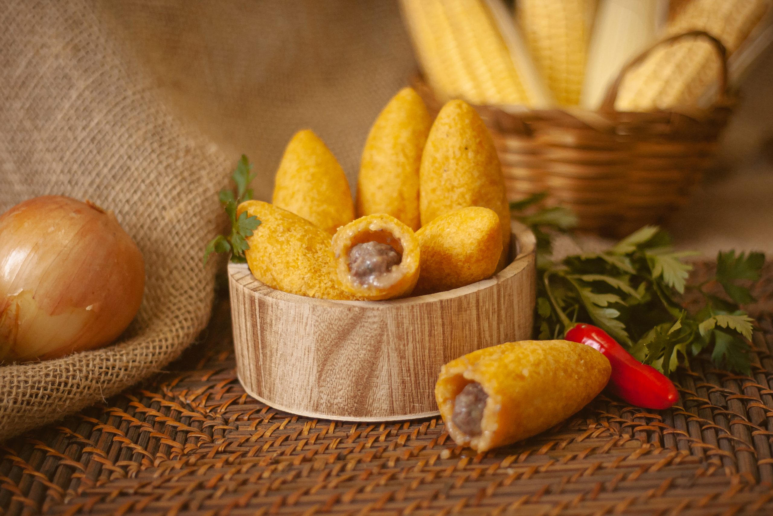 Uma cesta de madeira com diversos bolinhos caipiras de carne, com uma cesta de milho ao fundo, pimentas na mesa e uma cebola.
