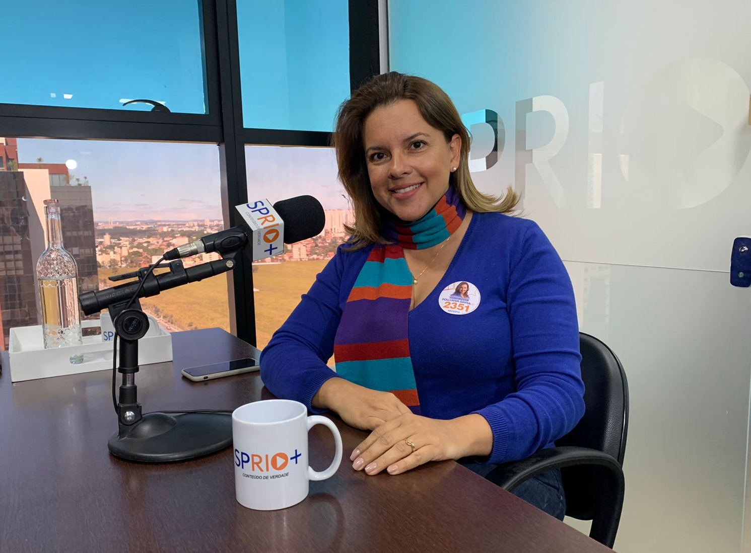 Pollyana Gama, ex-vereadora de Taubaté, sentada na mesa da SP RIO+, com uma caneca e um microfone, e a paisagem ao fundo.