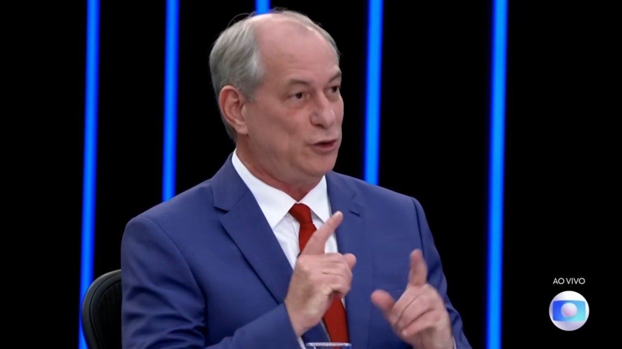 Ciro Gomes em entrevista ao JN. Ele veste terno na cor azul Bic, camisa branca e gravata vermelha. No frame, Ciro olha para em diagonal para os apresentadores e levanta os dois dedos indicadores na altura do peito.