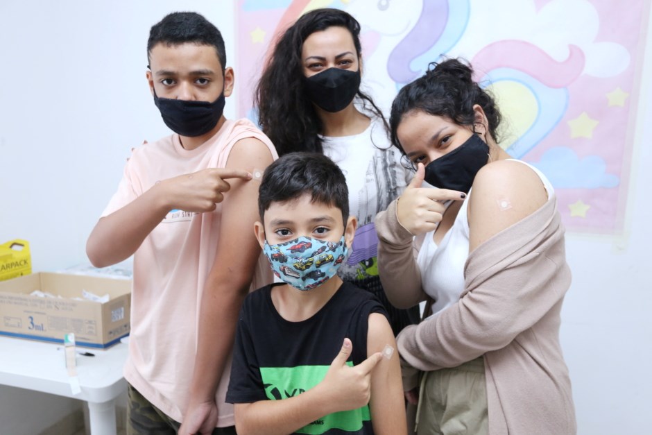 Grupo alvo da campanha de multivacinação, três crianças e jovens em Unidade de Saúde após receberem vacinas apontam para os braços em que receberam a imunização em gesto para incentivar vacinação. Junto delas também há uma mulher adulta atrás. Todos estão de máscara de proteção facial.