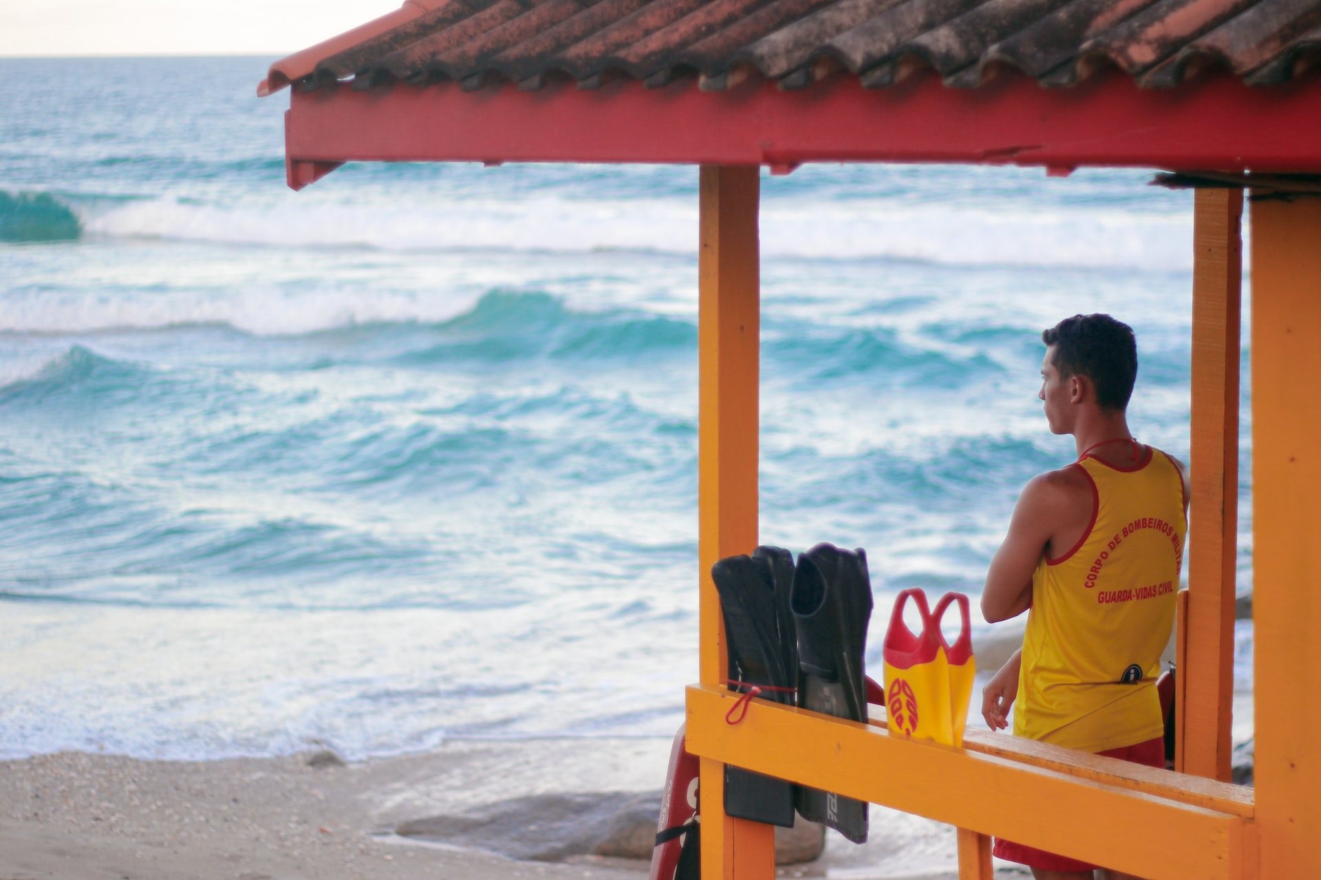 Salva-vidas no alto de posto de observação de uniforme amarelo e vermelho e braços cruzados, à esquerda da imagem, obsevando o mar.