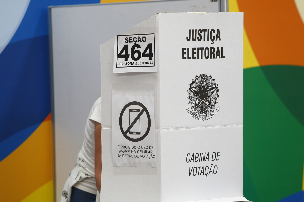 Eleitora de calça jeans e camiseta branca em cabine de votação registrando seu voto.