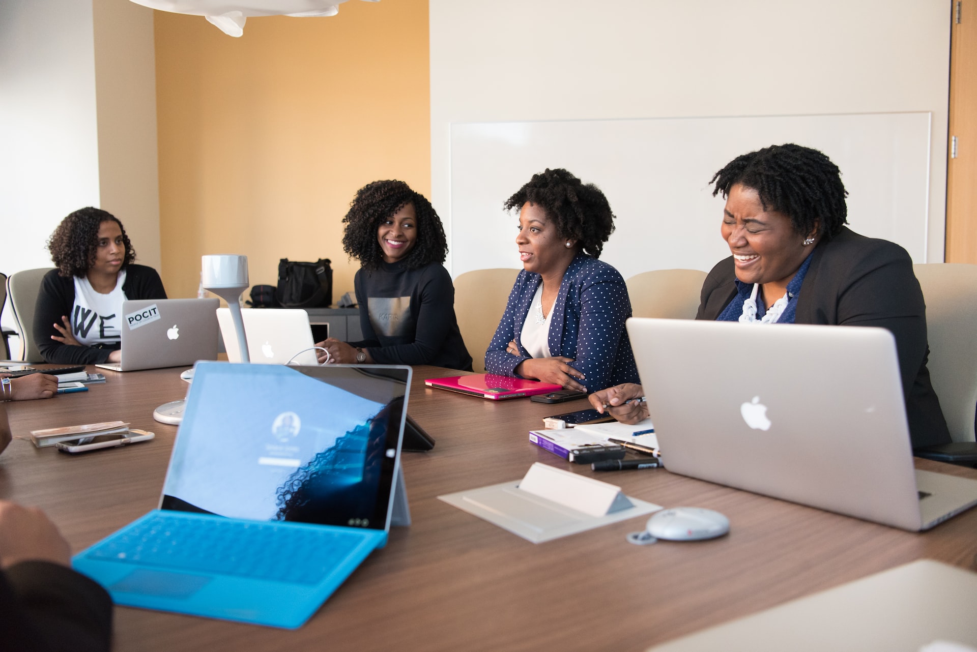 Quatro mulheres negras sentadas em mesa de escritório discutindo trabalho e negócios com bom humor. Cada uma delas tem um notebook em frente e na mesa há outros itens como canetas, smartphones e cadernos.