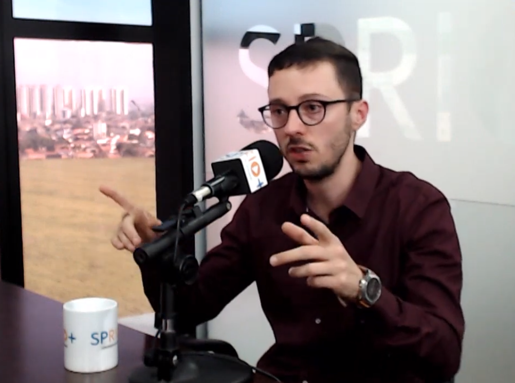 Fernando Ferreira, psicólogo em SJC, em entrevista à SP RIO+, falando ao microfone sobre a crise dos 25 anos, com uma caneca na mesa e a paisagem ao fundo.