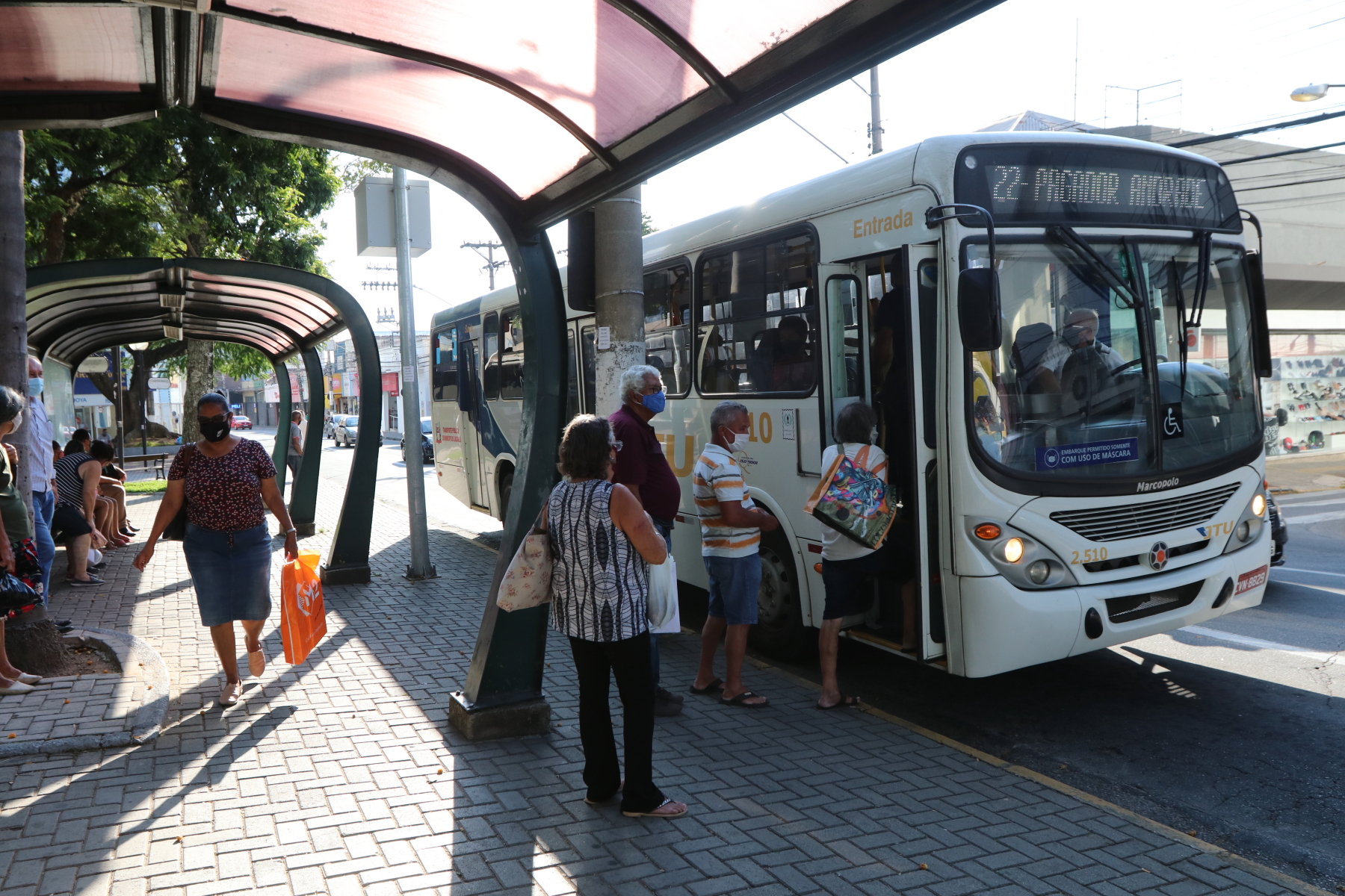 Ônibus em Jacareí parado em frente a ponto de ônibus para embarque de passageiros. Na fila esperando para entrar no ônibus estão quatro idosos. Também há mais pessoas esperando transporte sentadas no banco do ponto. 