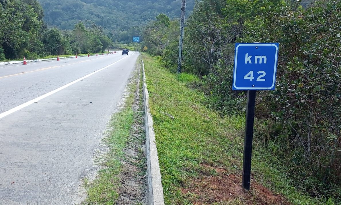 Foto de uma das placas implantadas na rodovia BR-101 (Rio-Santos), com uma estrada à esquerda e árvores à direita.
