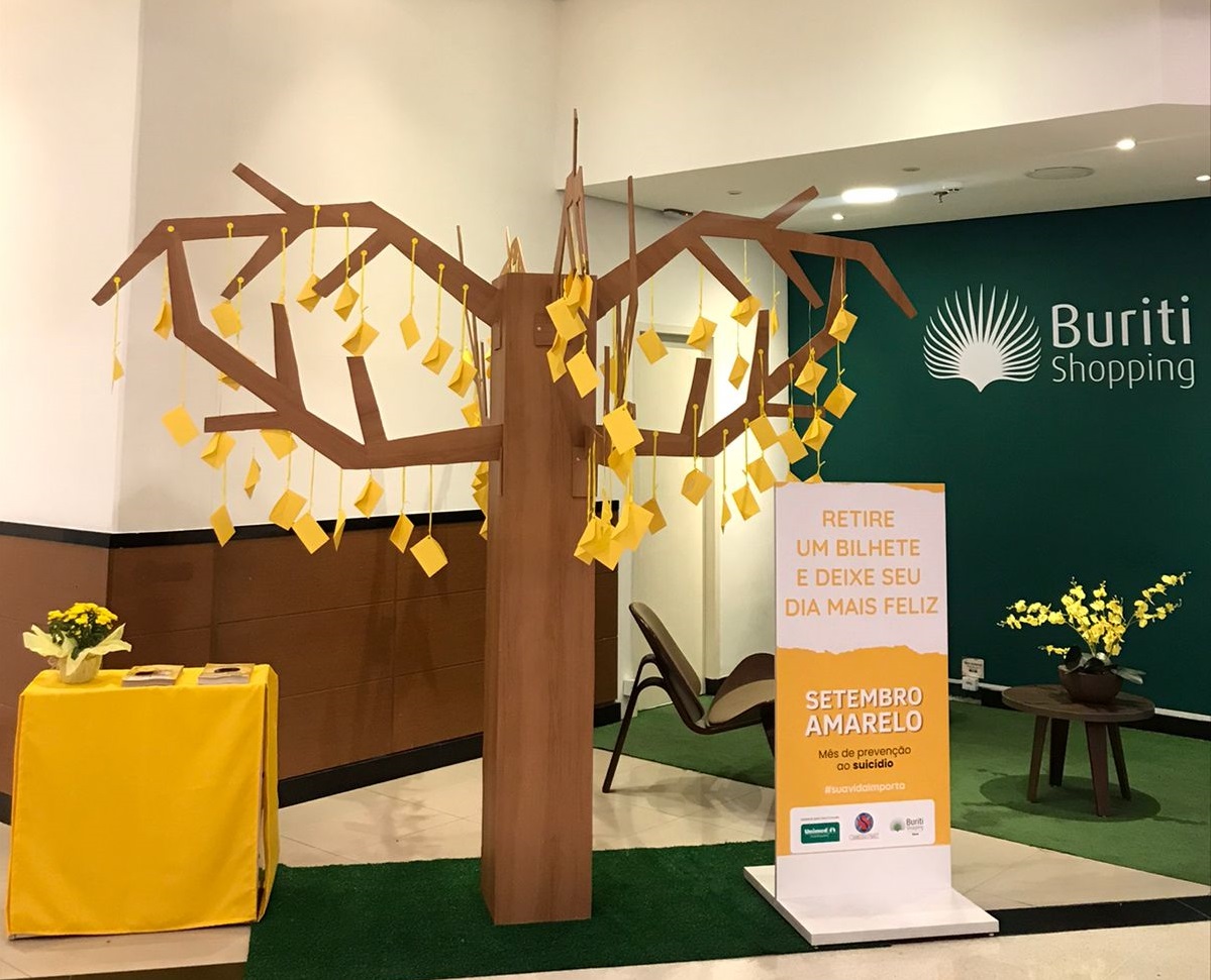 Ambiente decorado no Shopping de Guaratinguetá, com uma árvore decorativa no meio, papeis amarelos pendurados, uma mesa à esquerda, grama sintética e um cartaz à direita.