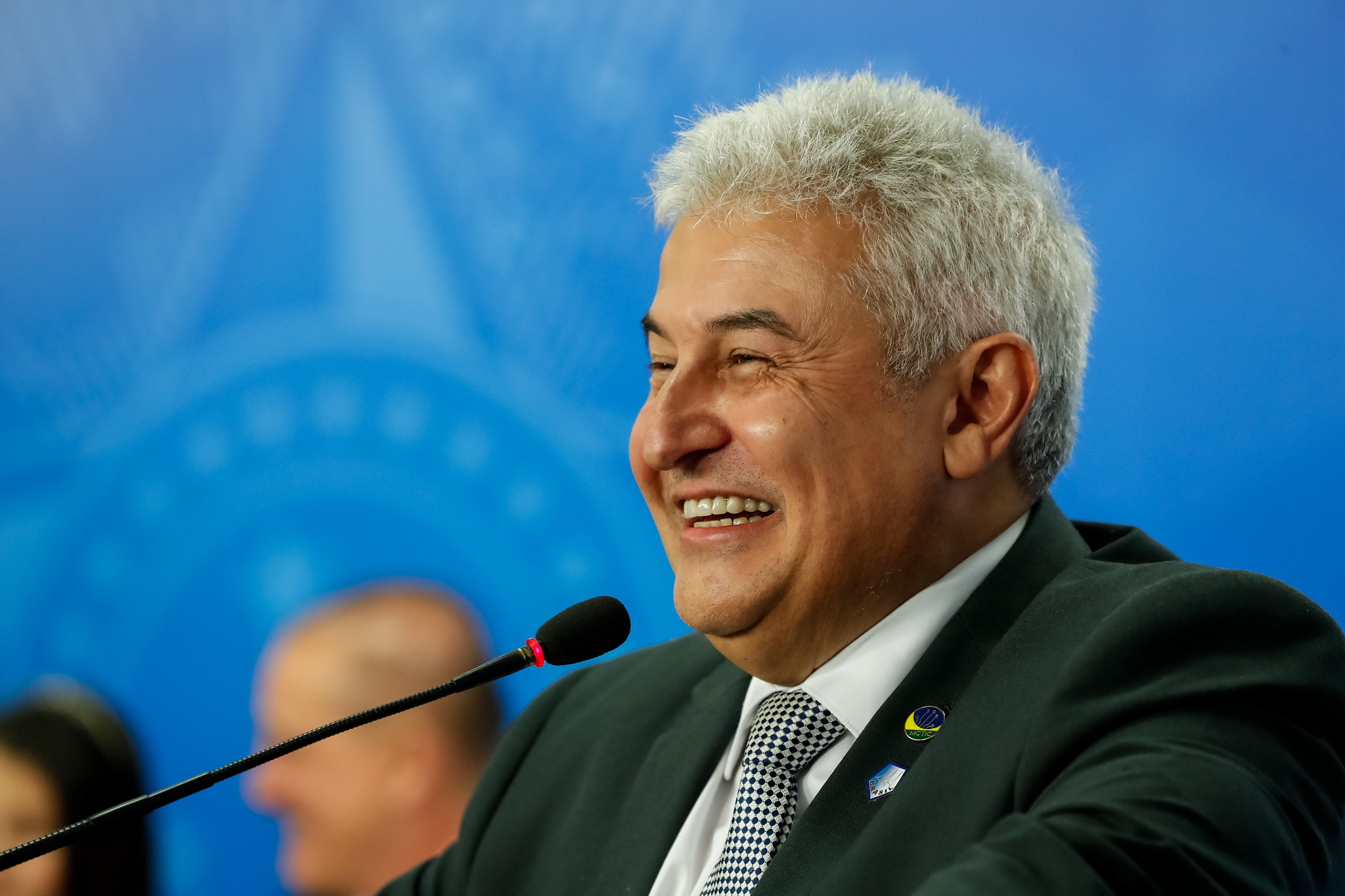 Astronauta Marcos Pontes, agora senador eleito, sorrindo para o lado esquerdo, com o fundo azul, falando no microfone, com um terno, camisa branca e gravata preta.