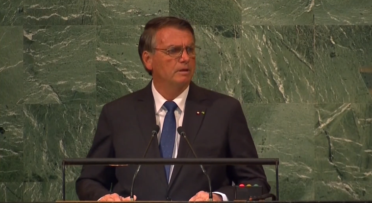 Bolsonaro com um terno preto, camisa branca e gravata azul, de óculos, olhando para o lado, em uma parede verde ao fundo, durante seu discurso na ONU.