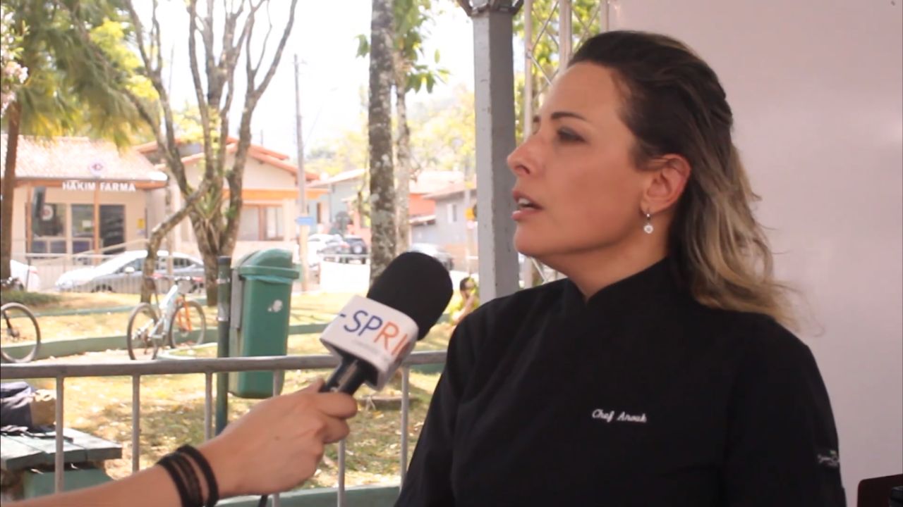 Mão segurando um microfone, apontando para a chef Anouk Migotto, com uma parede branco atrás, ao lado da praça.