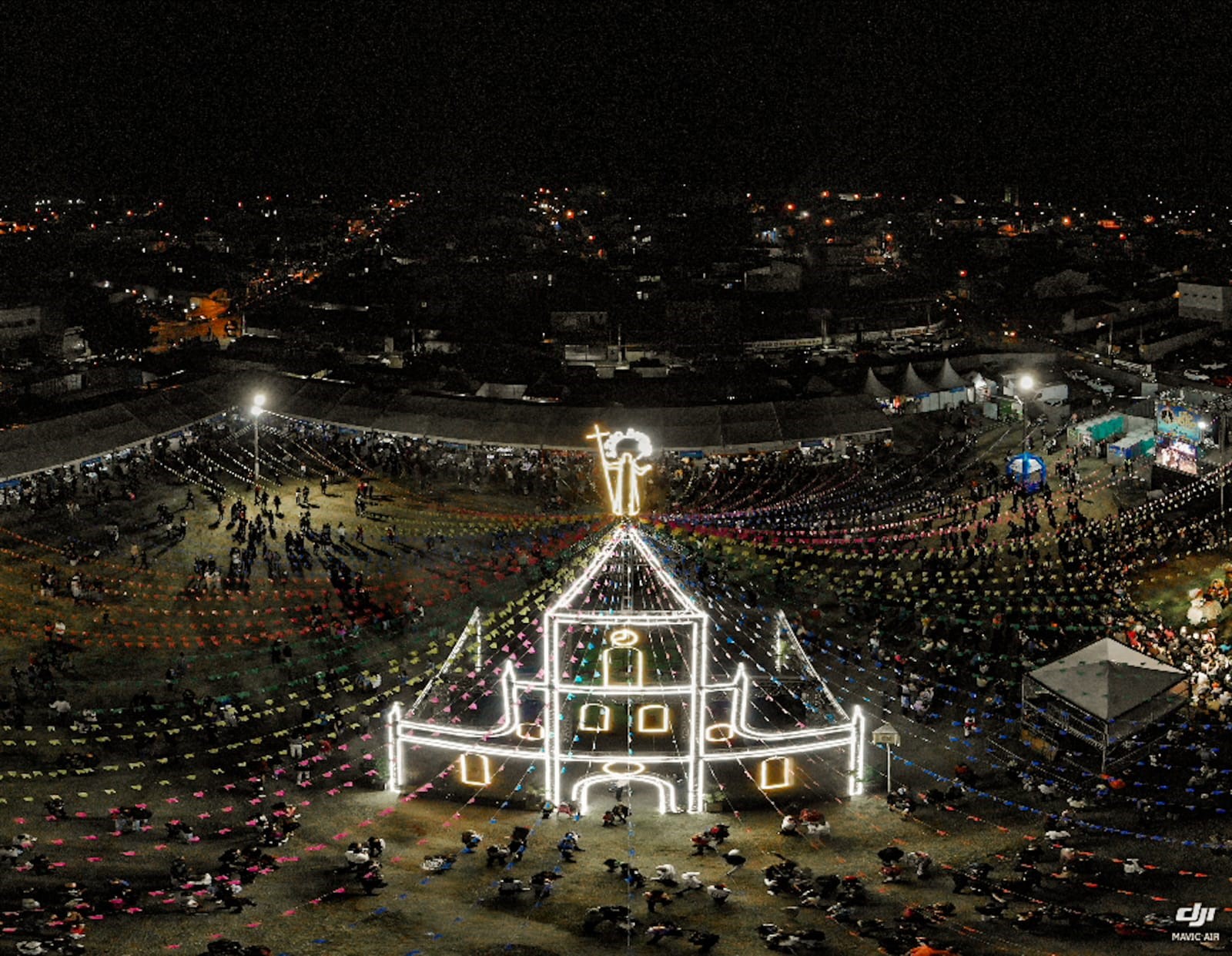 Vista aérea de uma Festa de João, com uma igreja iluminada, com bandeirinhas juninas coloridas em volta, diversas pessoas em volta e a cidade ao fundo. O local é um dos pontos turísticos de Caçapava.