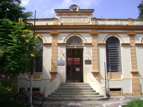 Fachada do Grupo Escolar Ruy Barbosa, em amarelo e branco, com árvores à esquerda, uma escada no meio dando para a calçada e uma bicicleta estacionada.