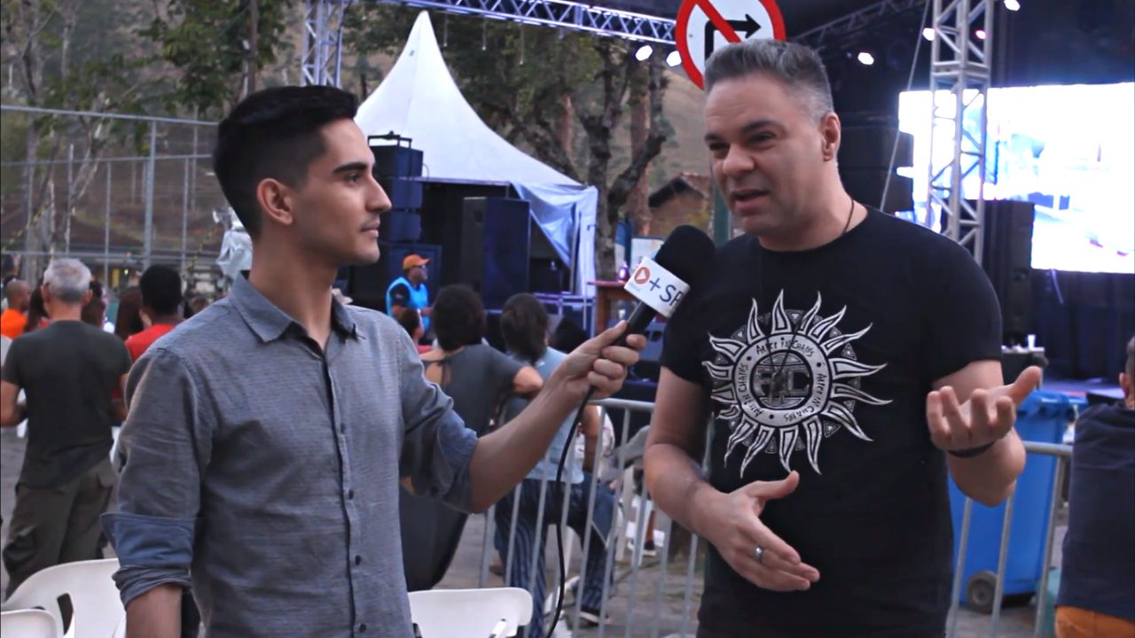 Matheus Andrade, jornalista, segurando um microfone do lado esquerdo, apontando para o entrevistado Peleco à direita, com o evento ao fundo, ao lado de um palco.
