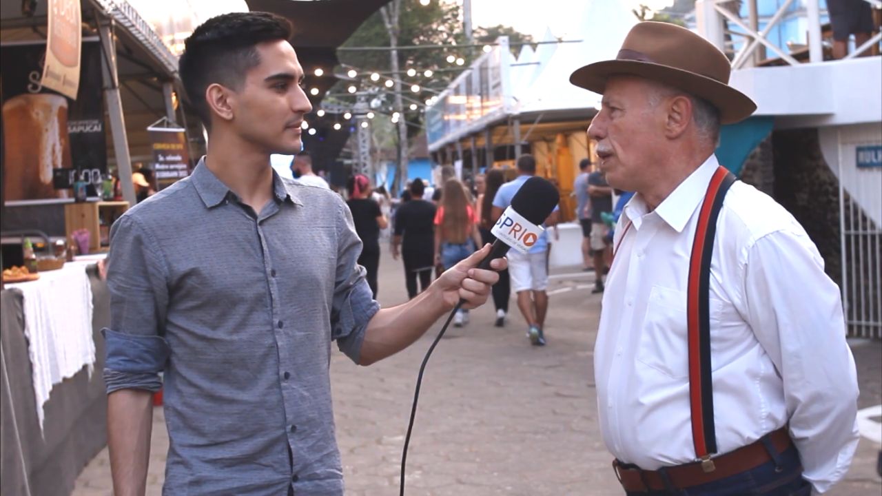 Jornalista Matheus Andrade segurando um mirofone, apontando para o entrevistado Carlito, à direita, que veste uma camisa branca, um suspensório vermelho e um chapéu, com o evento ao fundo.