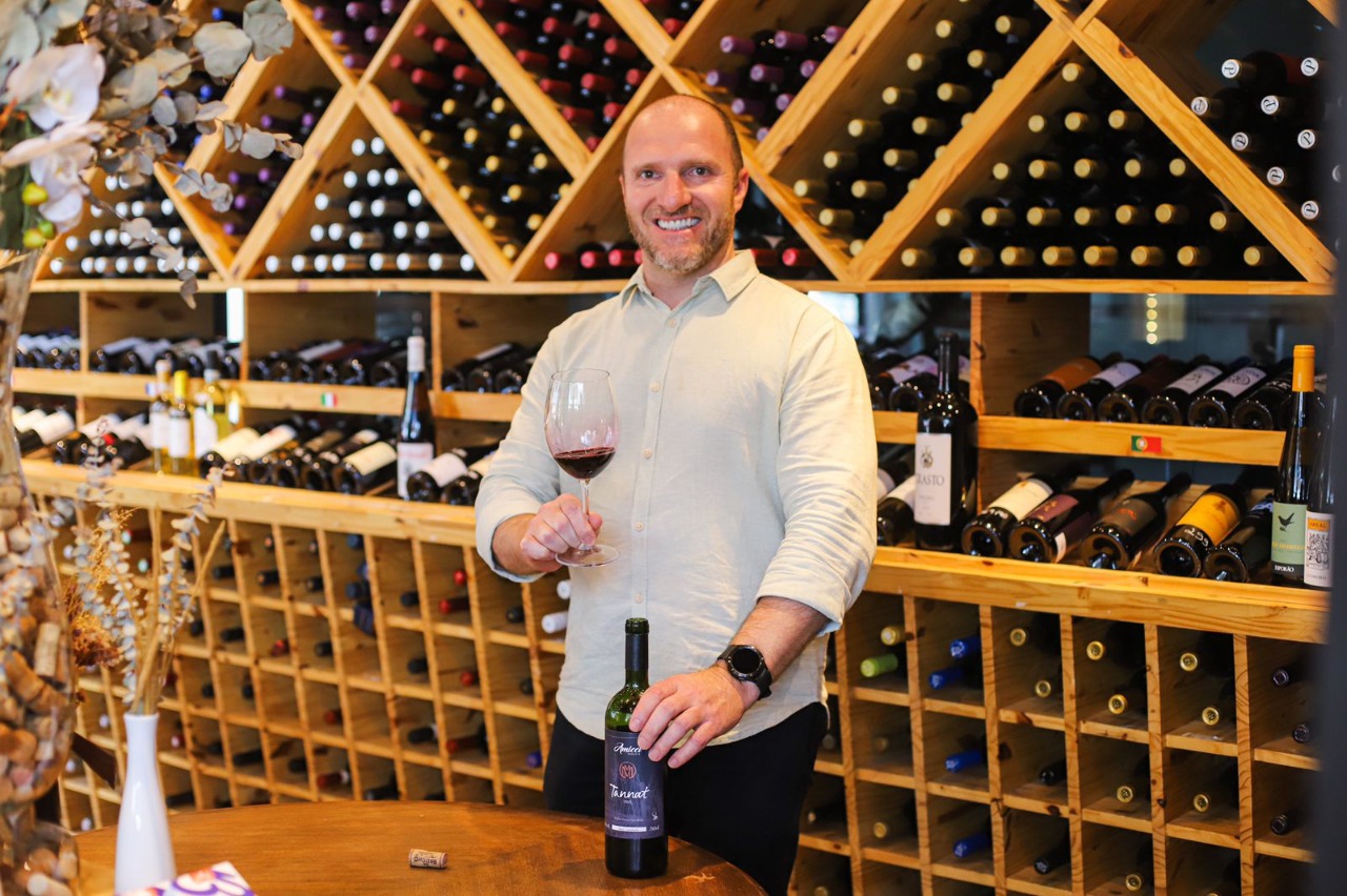 Marcelo de Moraes com uma taça de vinho na mão em uma adega repleta de variados vinhos. Ele está sorrindo para a câmera.