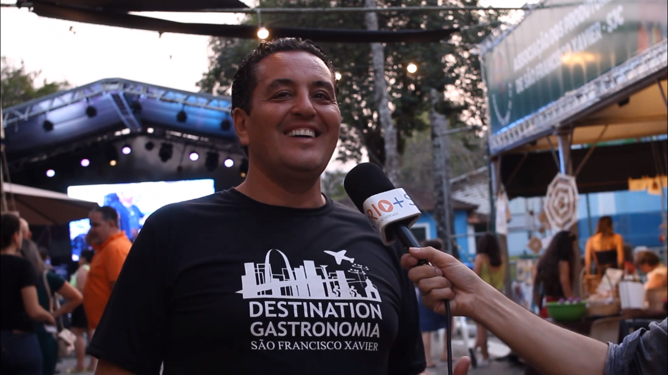 Mão segurando um microfone e apontando para o entrevistado Mauricio Guisard, à esquerda, vestindo uma camisa preta, sorrindo para a câmera, com o palco do Destination ao fundo.