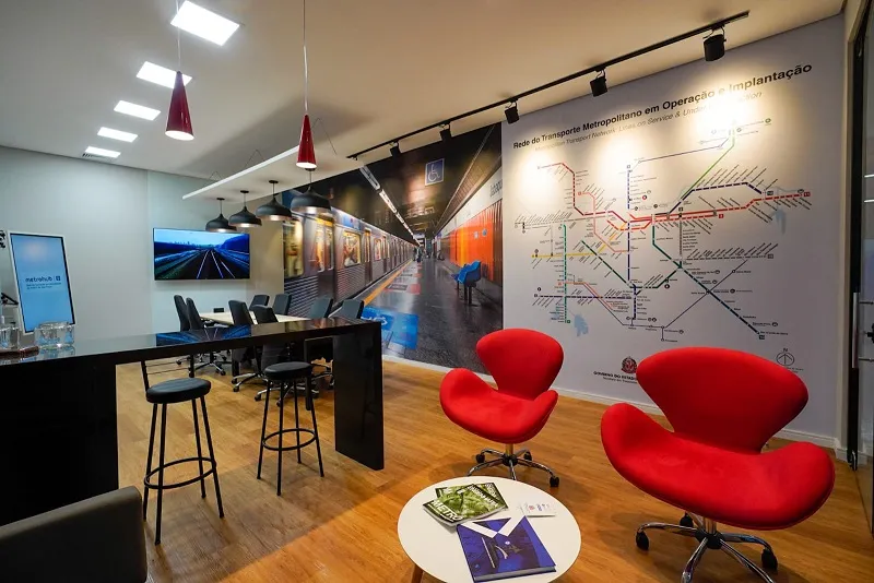 Sala da startup Metrohub, do Metrô de SP, que foi implantada no Parque Tecnológico, com cadeiras vermelhas, uma placa branca com as linhas do metrô, chão de madeira, mesa e teto branco, com luminárias brancas.