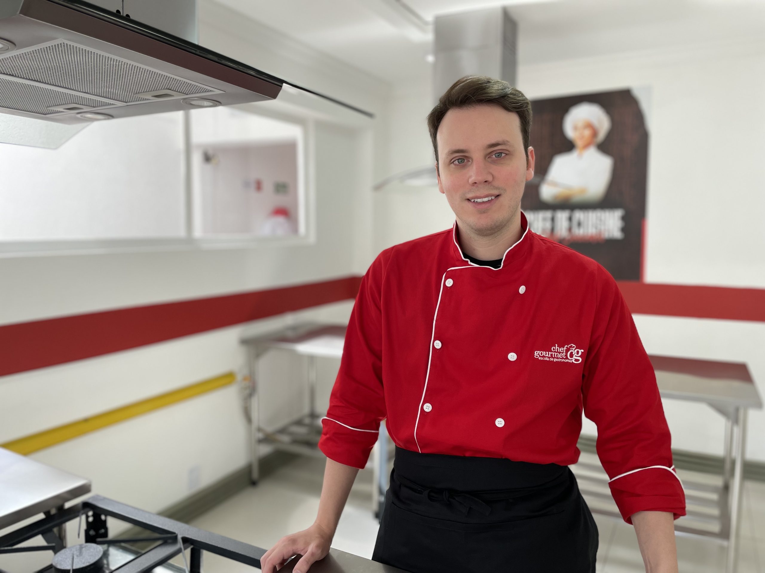 Rafael Ferro, diretor da Escola de Gastronomia Chef Gourmet Taubaté, olhando para a câmera, com um uniforme vermelho, calça preta e a cozinha ao fundo.