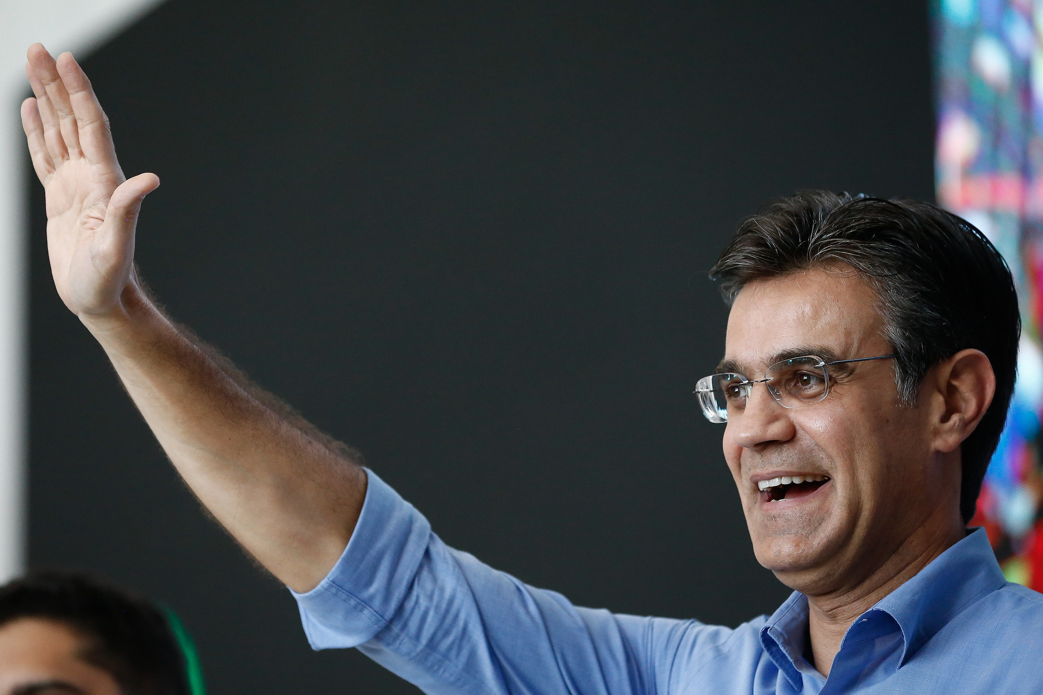 Governador Rodrigo Farcia com uma camisa azul, sorrindo, acenando para a esquerda, com um fundo escuro.