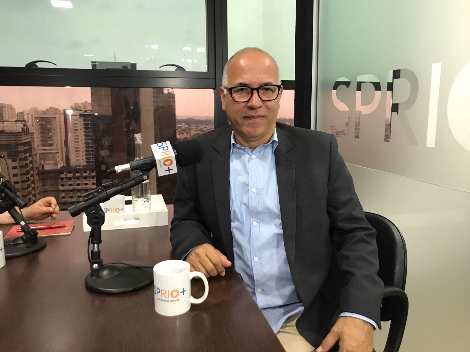Professor Agliberto Chagas nos estúdios da SP RIO+. Ele é homem, branco, careca e apresenta semblante neutro. Agliberto aparece na foto sentado, da cintura para cima, e veste camisa social azul clara e blazer cinza escuro.