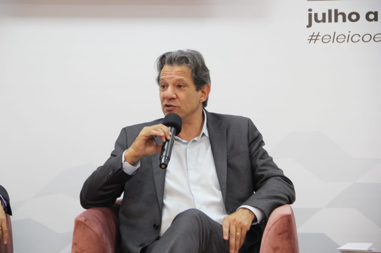 Fernando Haddad durante participação em sabatina São Paulo em Debate, realizada em São José dos Campos. O candidato ao governo de SP aparece do tronco para cima, sentado e com as pernas cruzadas. Ele veste camisa social branca, blazer e calça cinza e fala em microfone.