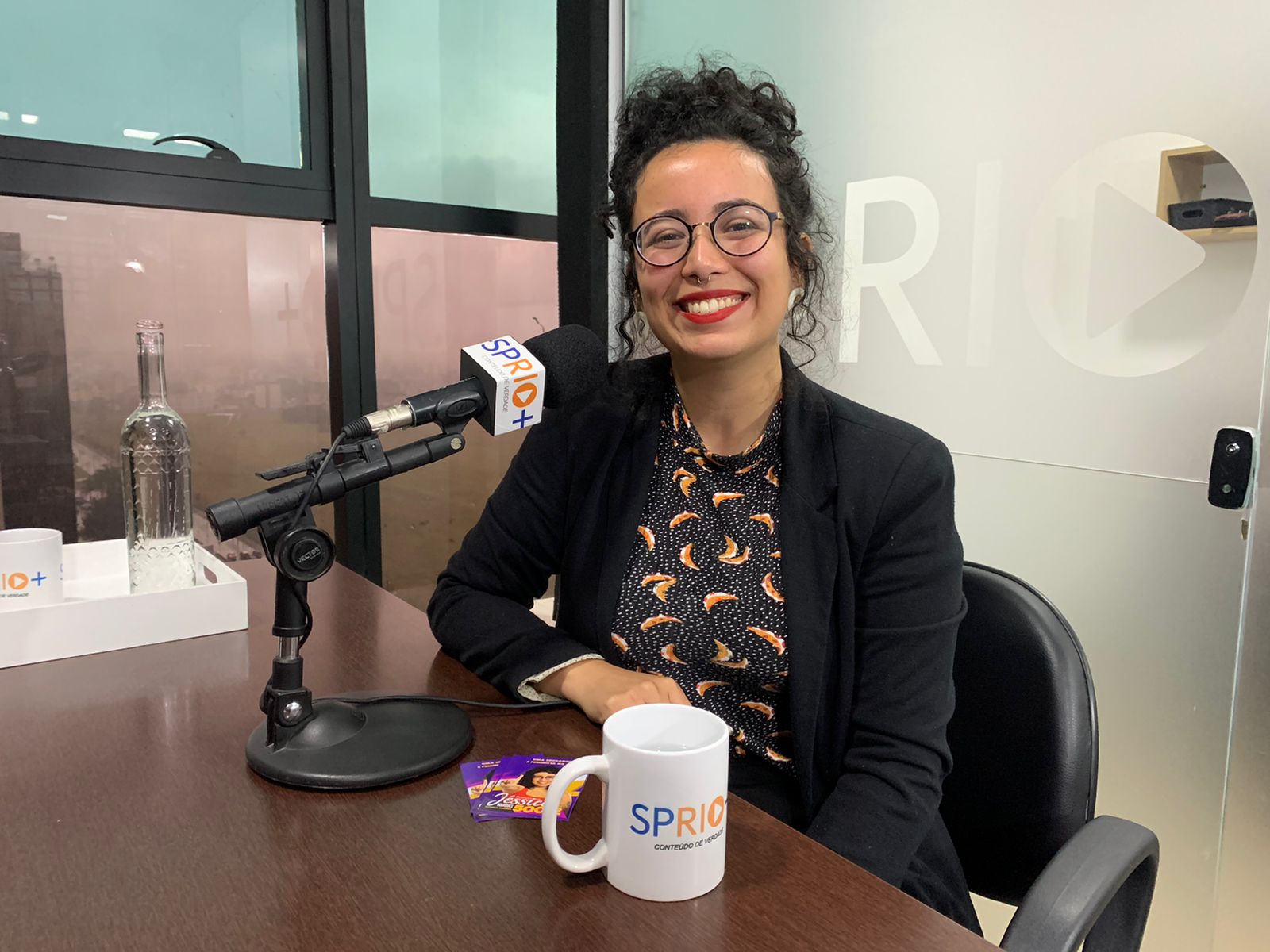 Candidata a deputada estadual Jéssica Marques nos estúdio da SP RIO+ após entrevista. Ela aparece sentada, do peito para cima e sorrindo. Ela é branca, usa batom vermelhos, brincos brancos nas orelhas e possui cabelo ondulado etem cabelo ondulado, preso no momento. Jéssica ainda veste blazer preto com camisa social preta estampada com pequenas bolinhas brancas e outros padrões.