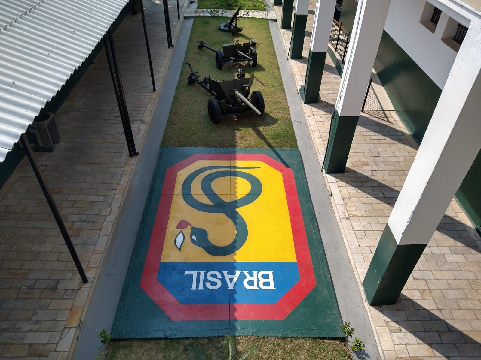 Vista aérea de um corredor do 6º Batalhão de Infantaria Leve, em Caçapava, com um jardim no meio, uma placa escrito Brasil, pilastras ao lado nas cores verdes e brancas, além de veículos militares parados.