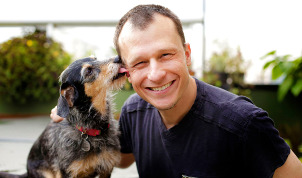 Alexandre Rossi, adestrador e especialista em comportamento pets, ao lado de cachorro, que o lambe.