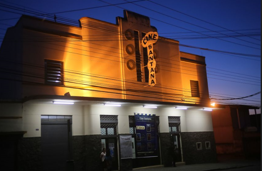 Fachada do Cine Santana, à noite, com uma placa indicando o nome do local, e as portas abaixo.