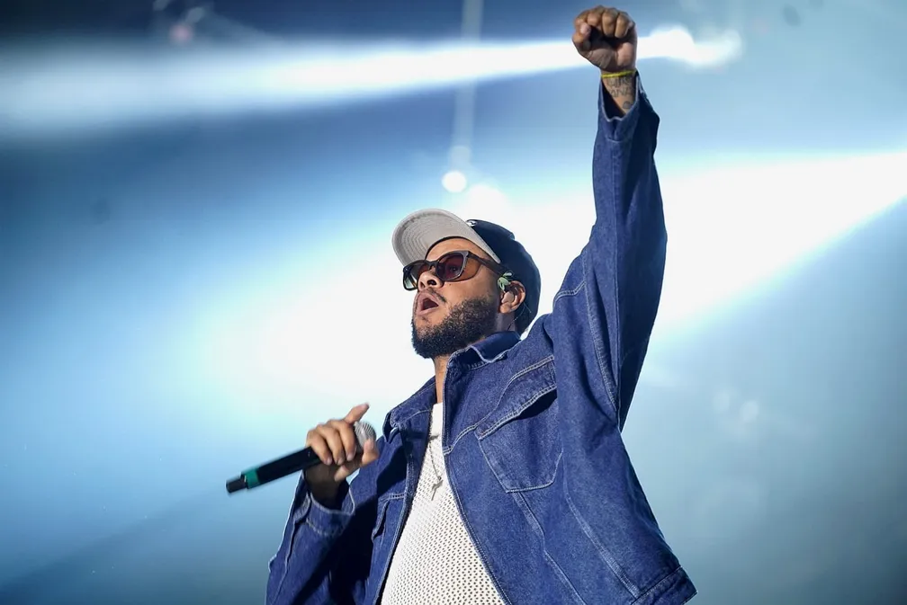 Emicida alfinetou Bolsonaro no Rock in Rio, vestindo uma jaqueta jeans azul, um boné preto, óculos de sol e com o braço para cima.