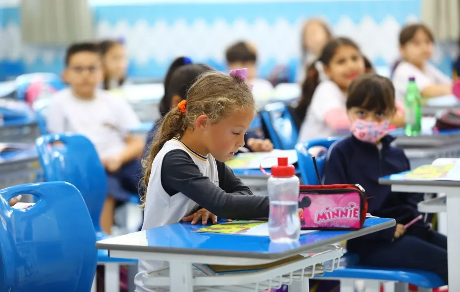 Alunos da rede pública de SJC, com uma garrafinha na mesa azul, um estojo da Minnie e alunos ao fundo. 