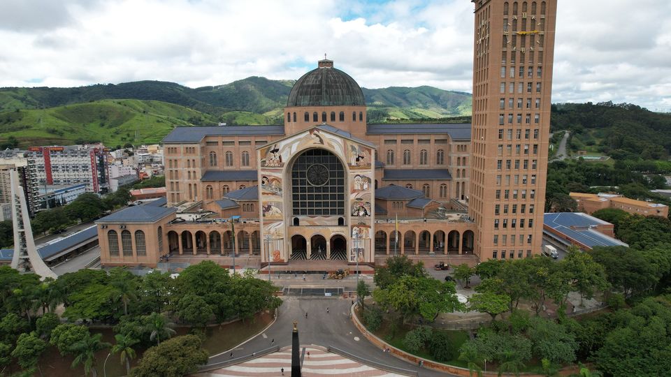 Vista aérea do Santuário Nacional de Aparecida, com céu azul, construção no meio e área verde na frente.