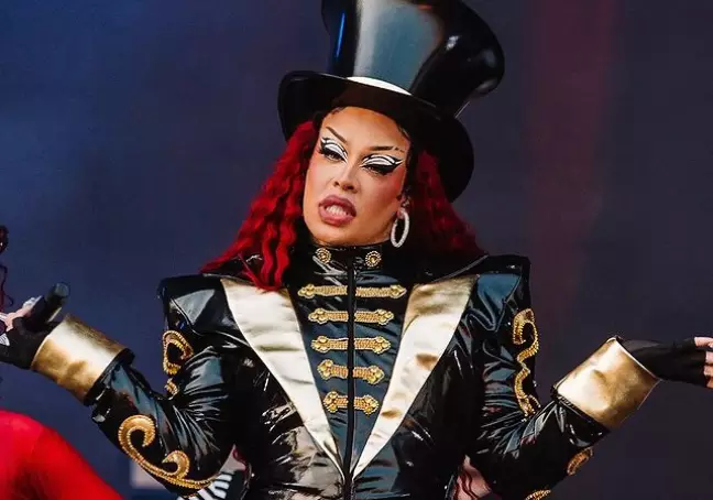 Gloria Groove com o cabelo vermelho, um chapéu preto, roupas pretas, com os braços levantados para o lado.