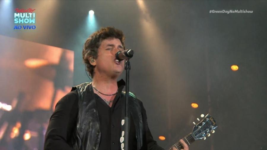Vocalista do Green Day cantando no Rock in Rio, com roupa preta, violão nos braços, e fundo preto.
